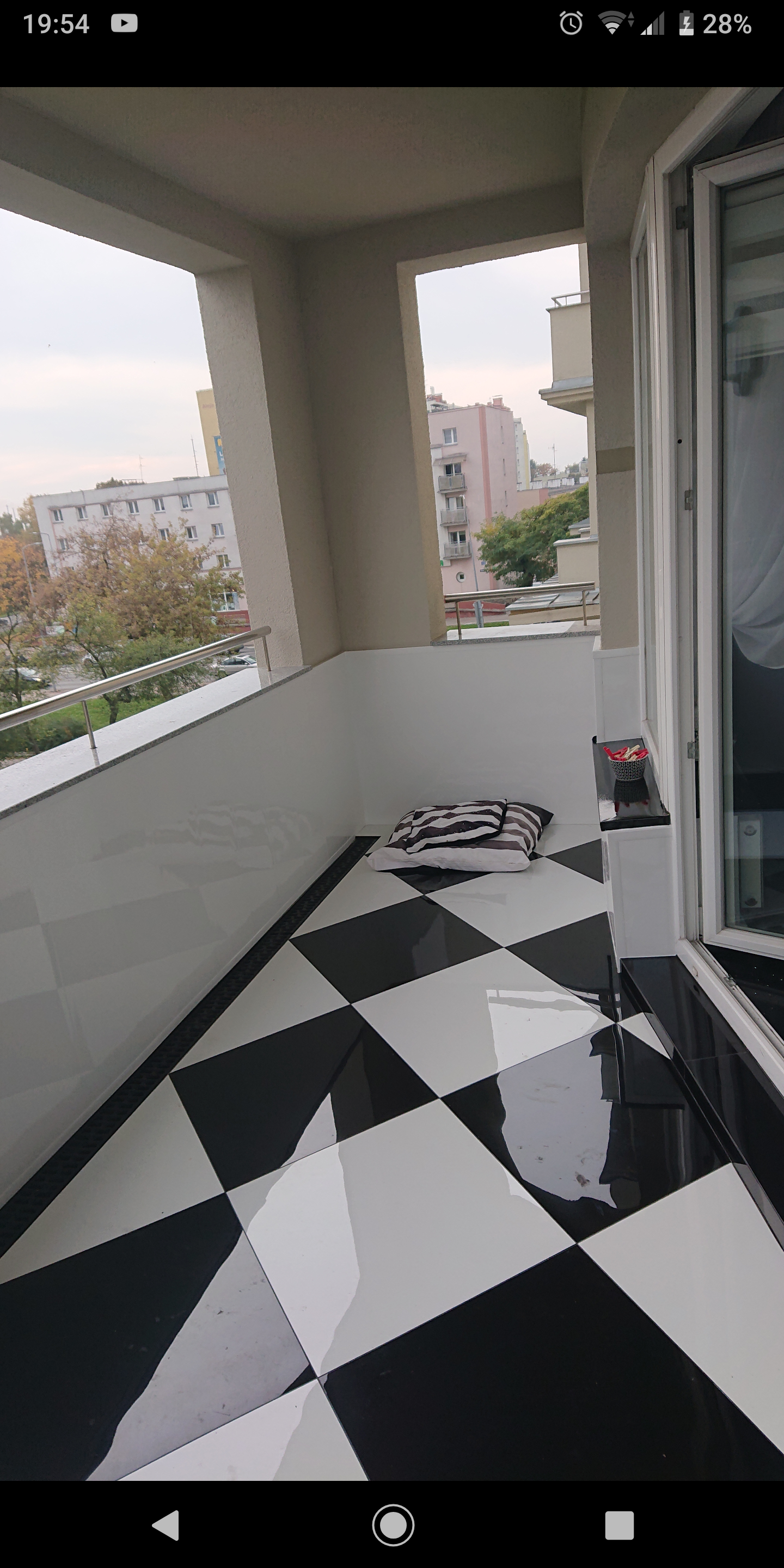 Balkon z podłogą w czarno-białą szachownicę, widok na miasto w tle, poduszki na podłodze.