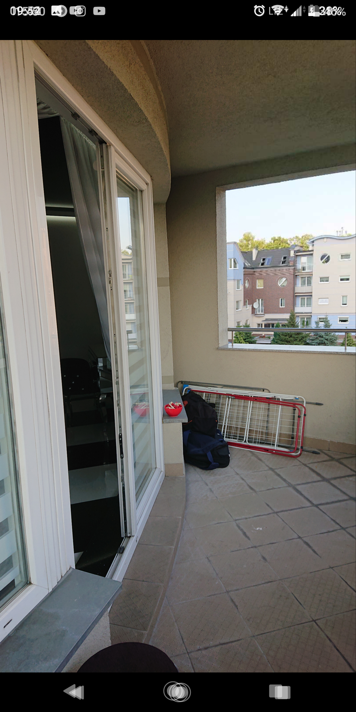 Balkon z otwartymi drzwiami balkonowymi, na podłodze widoczna suszarka na pranie, torba podróżna i miska z owocami, w tle budynki mieszkalne.