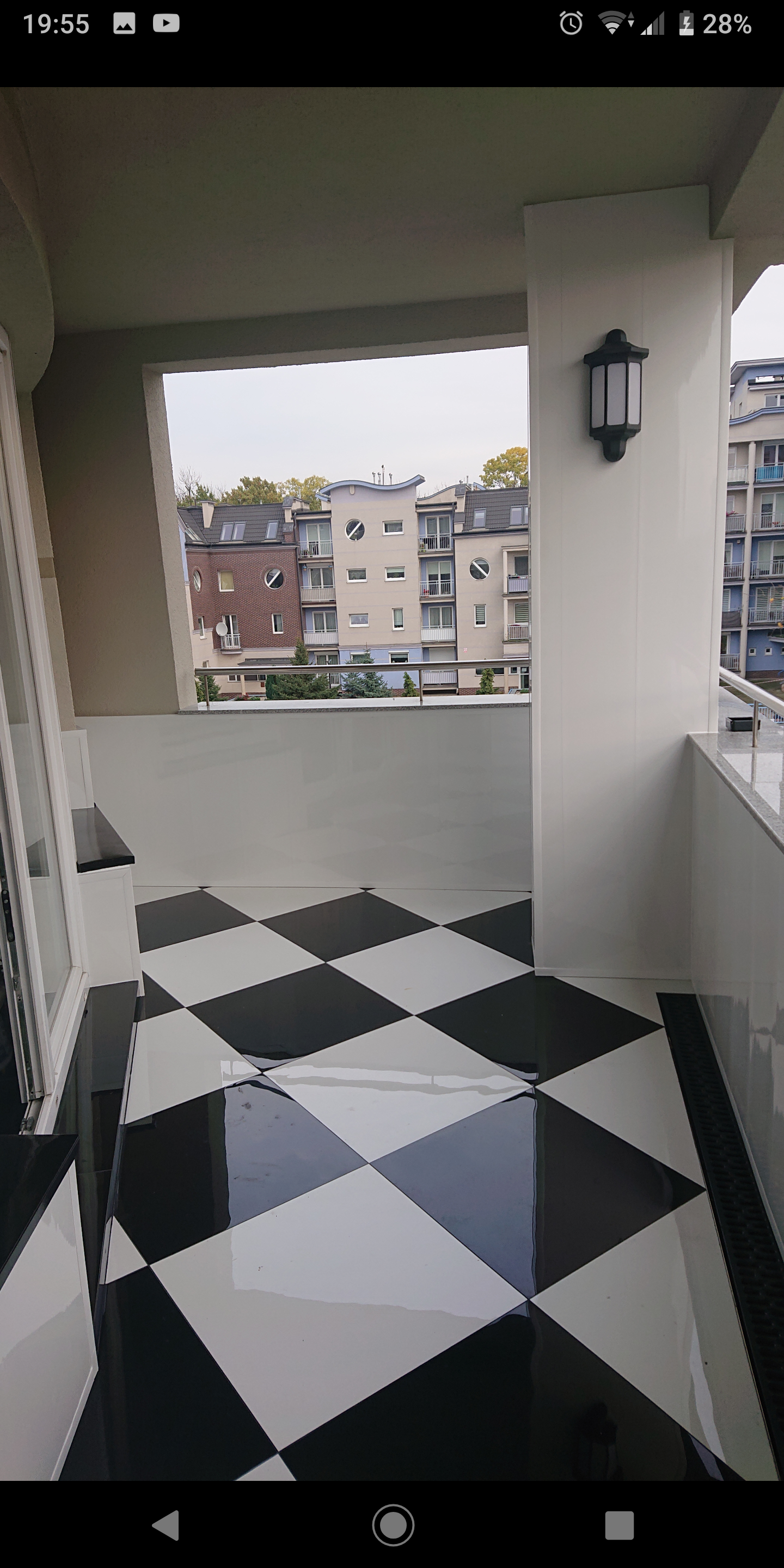 Błyszcząca, czarno-biała podłoga w szachownicę na balkonie z widokiem na budynki mieszkalne, oświetlona zewnętrzną lampą ścienną.