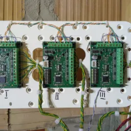 Trzy zielone moduły sterujące Satel INT-E V1.1 zamontowane na białej perforowanej płycie z numerami I, II, III, podłączone kolorowymi przewodami do listew zaciskowych, widoczne elementy instalacji...