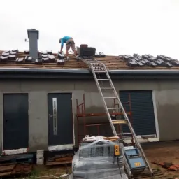 Pracownik w niebieskiej koszulce układa dachówkę na drewnianej konstrukcji dachu, używając wózka wciąganego po drabinie; widoczny komin i surowa elewacja domu z szarymi drzwiami i roletą.