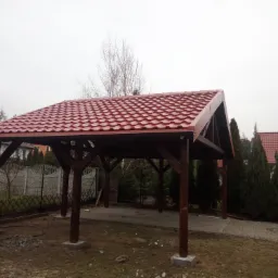 Drewniana altana z czerwonym dachem z blachodachówki, widoczna z zewnątrz, na tle szarego nieba i ogrodzenia.