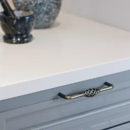 Szare szuflady kuchenne z dekoracyjnymi metalowymi uchwytami w stylu rustykalnym, na blacie czajnik w plamki i moździerz z pistlem.