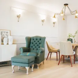 Turkusowy fotel typu uszak z podnóżkiem w jasnym salonie z okrągłym stołem, ozdobną lampą i białymi meblami, dekoracje w postaci rzeźby Buddy i wazonów.