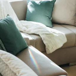 Beżowa sofa z dekoracyjnymi poduszkami w odcieniach zieleni i beżu oraz puszystym białym pledem, oświetlona promieniami słońca tworzącymi tęczę.