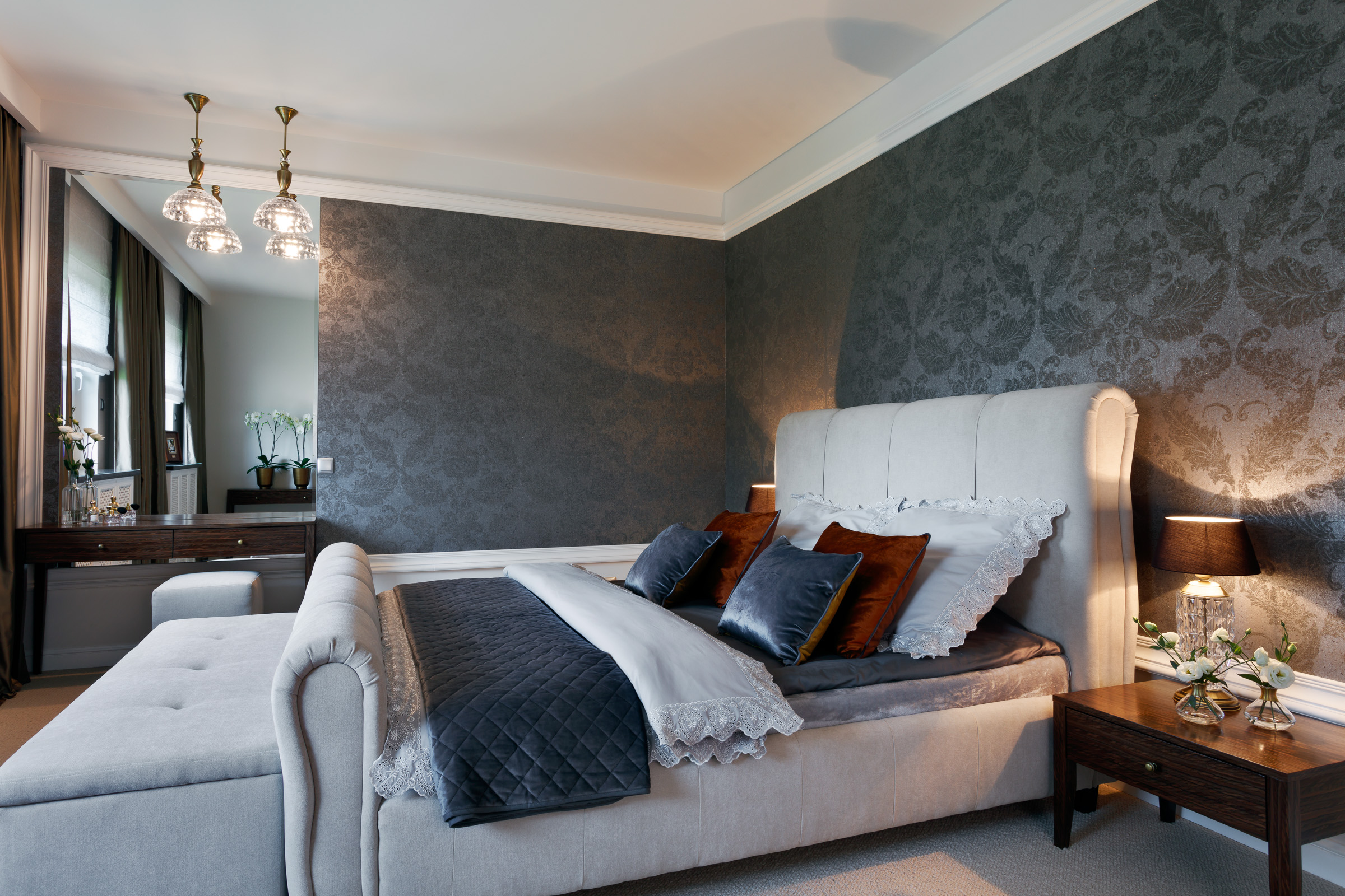 Sypialnia w tonacji szarości z łóżkiem typu king-size z tapicerowanym wezgłowiem, wzorzystą tapetą, stolikiem nocnym z lampką i kwiatami oraz toaletką z lustrem w tle.