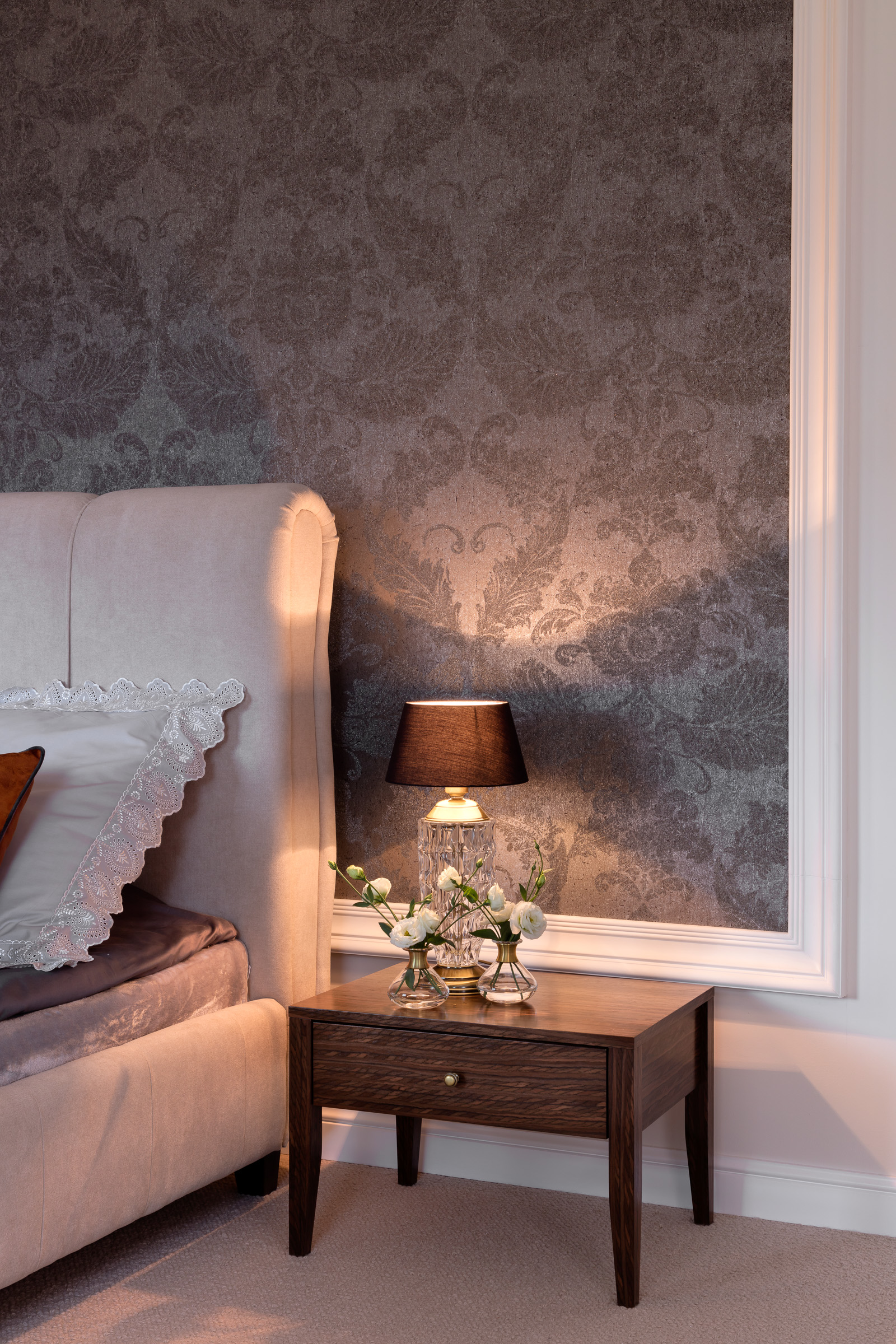 Sypialnia z tapetą w kwiatowe wzory, beżowym zagłówkiem łóżka, drewnianą szafką nocną z kwiatami w wazonach i kryształową lampą z czarnym abażurem.