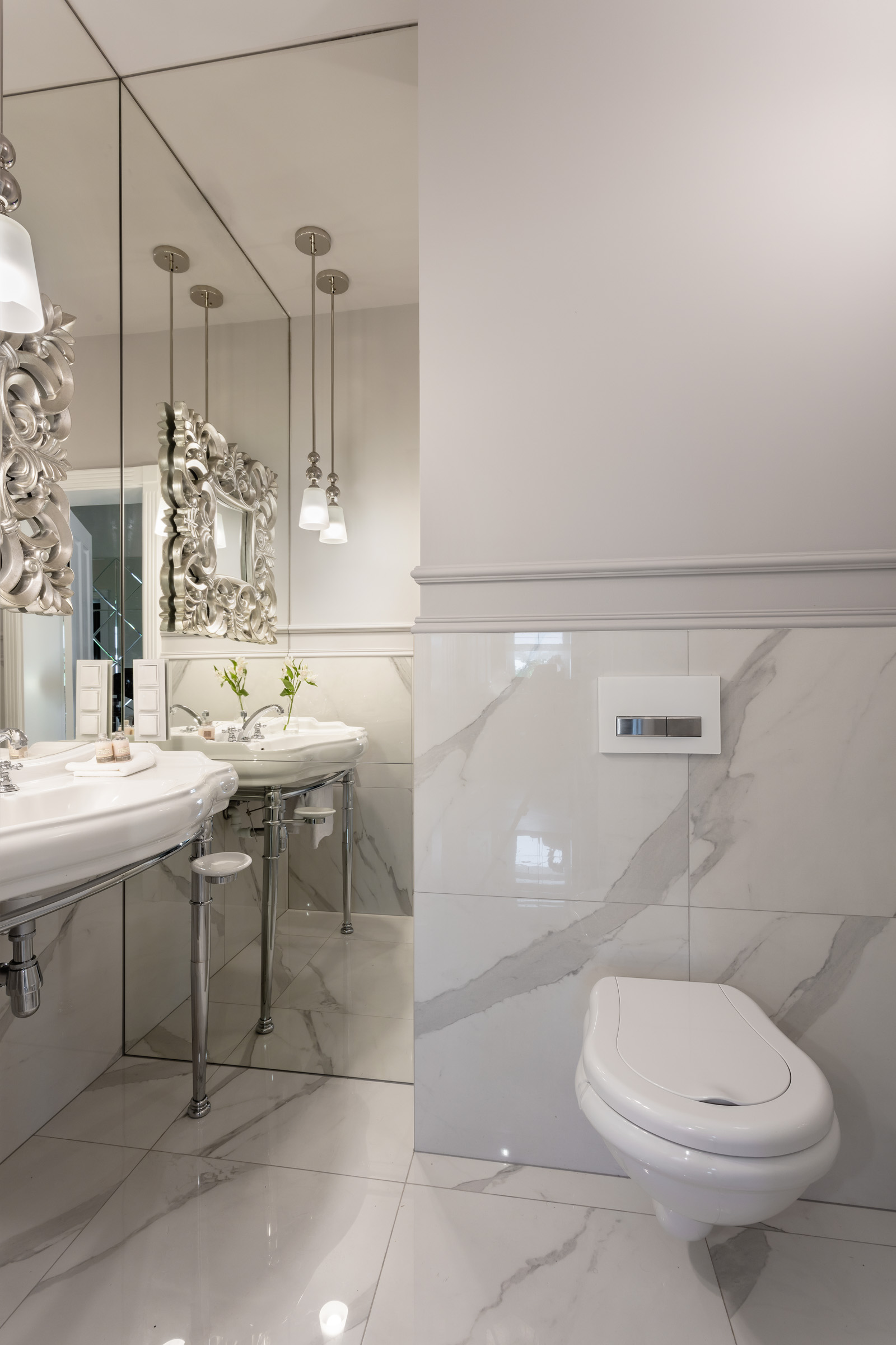 Elegancka łazienka w odcieniach bieli i szarości z marmurowymi płytkami, podwieszana toaleta, dwie umywalki na chromowanych nogach z lustrami w ozdobnych srebrnych ramach i wiszące lampy.