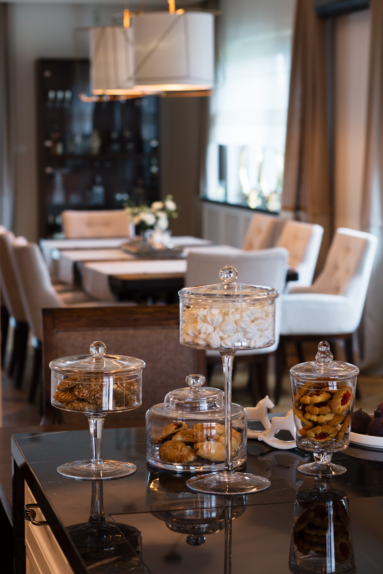 Elegancka aranżacja stołu z szklanymi pojemnikami na nóżkach wypełnionymi ciastkami i bezami, w tle stół z krzesłami i przeszklona witryna.
