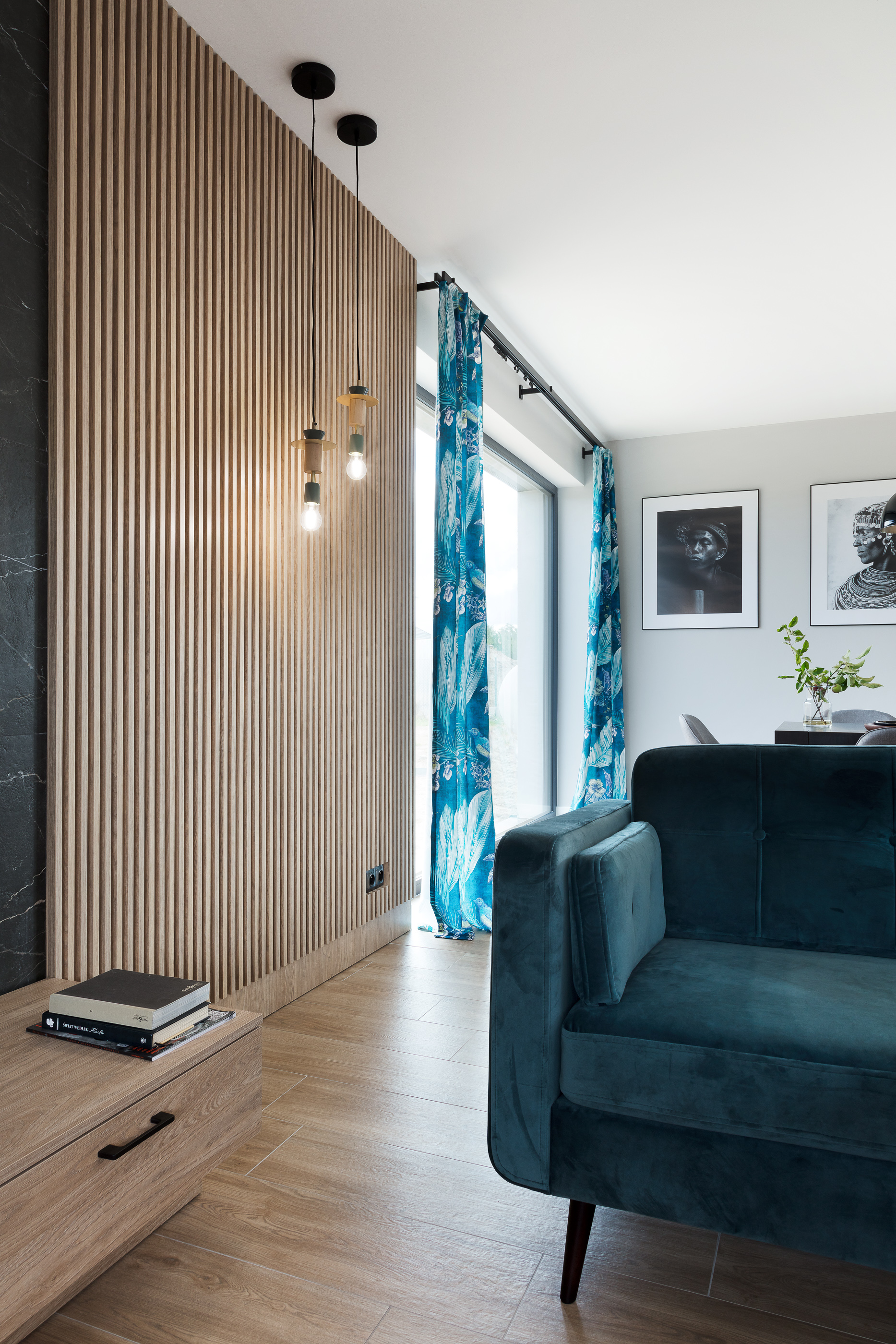 Salon z panelem ściennym z drewnianych listewek, turkusową sofą i zasłonami w kwiatowe wzory, uzupełniony o wiszące lampy i czarno-białe portrety na ścianie.
