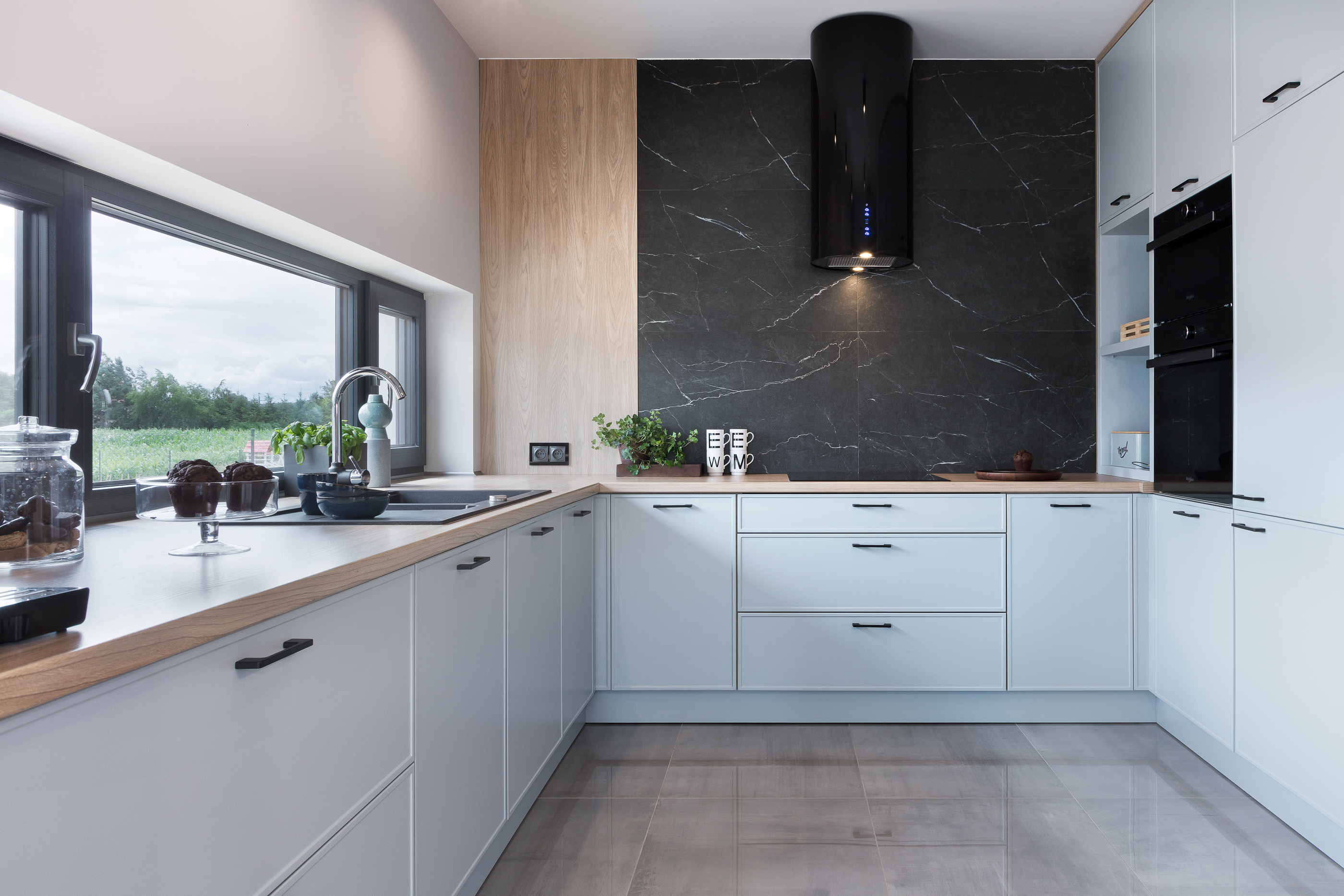 Jasna kuchnia z drewnianym blatem, czarnym okapem nad płytą, z widokiem na zielone pola za oknem, oraz z elementami dekoracyjnymi jak muffiny i zioła.