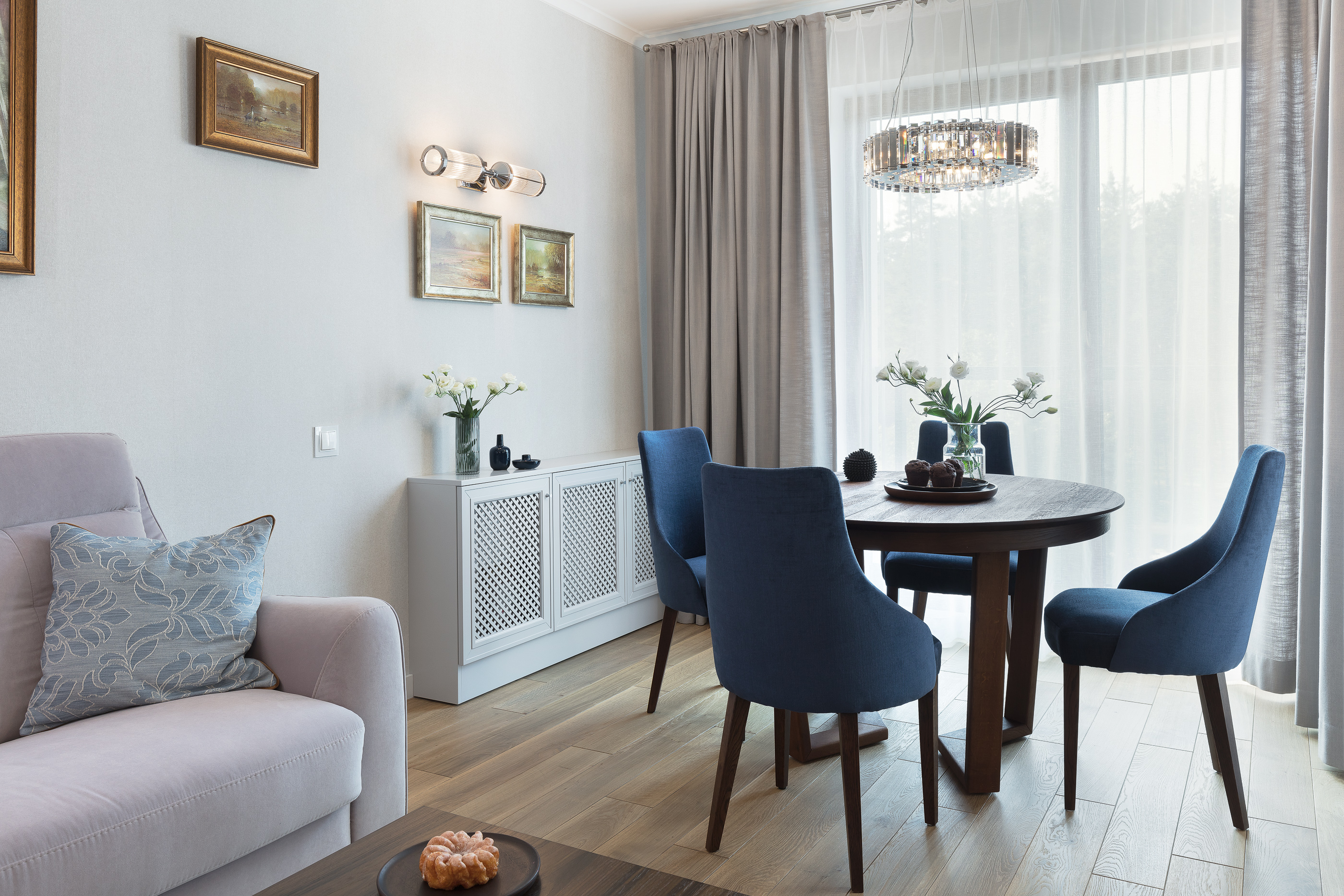 Jasny salon z okrągłym stołem, niebieskimi krzesłami, sofą w odcieniach fioletu i białymi kwiatami w wazonie, oświetlony nowoczesną lampą sufitową.