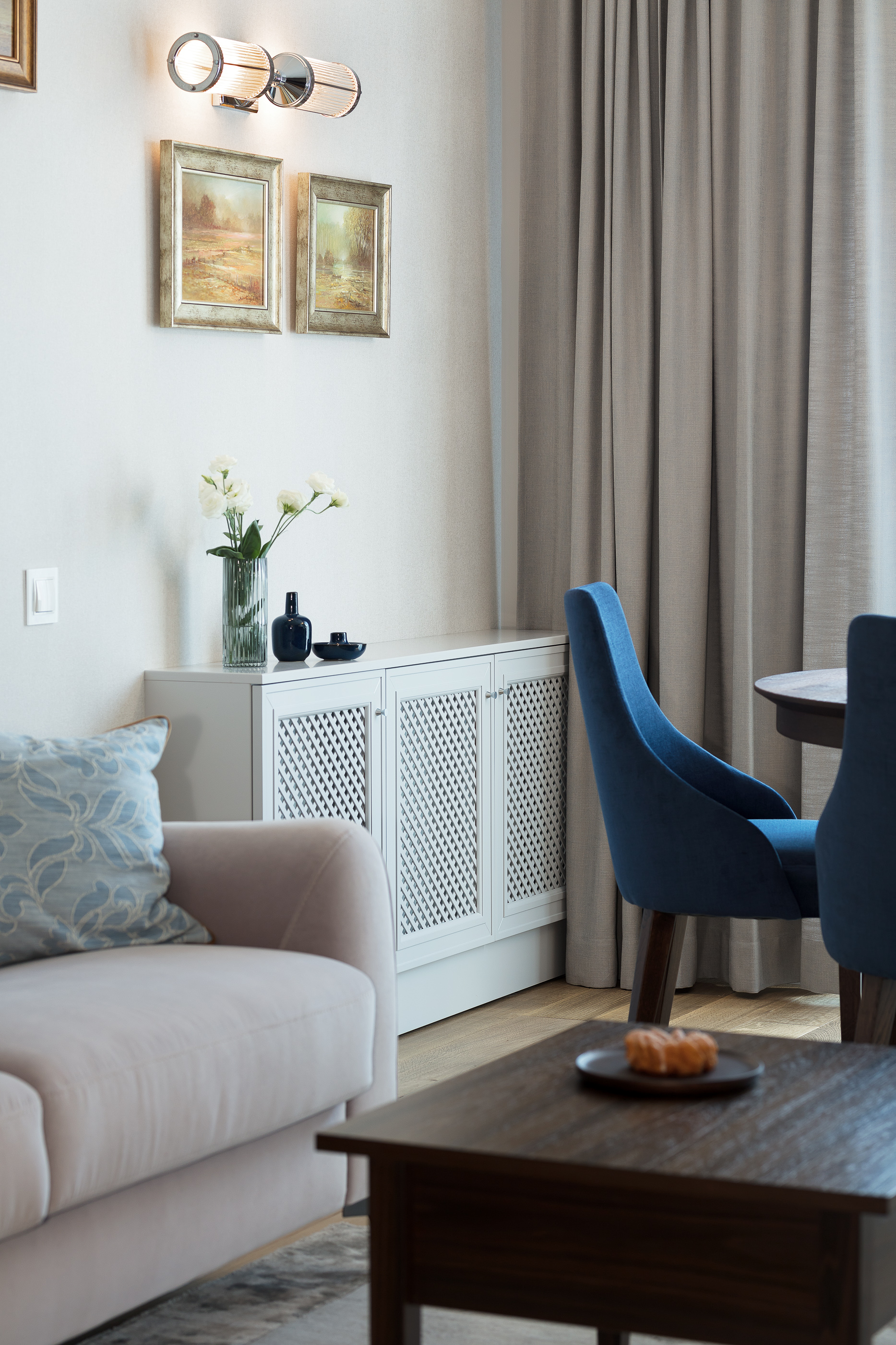 Jasny salon z beżową sofą z dekoracyjną poduszką, białym kredensem z ażurowymi drzwiczkami, wazonem z białymi kwiatami, granatowymi detalami dekoracyjnymi, ciemnym stolikiem kawowym z przekąską...