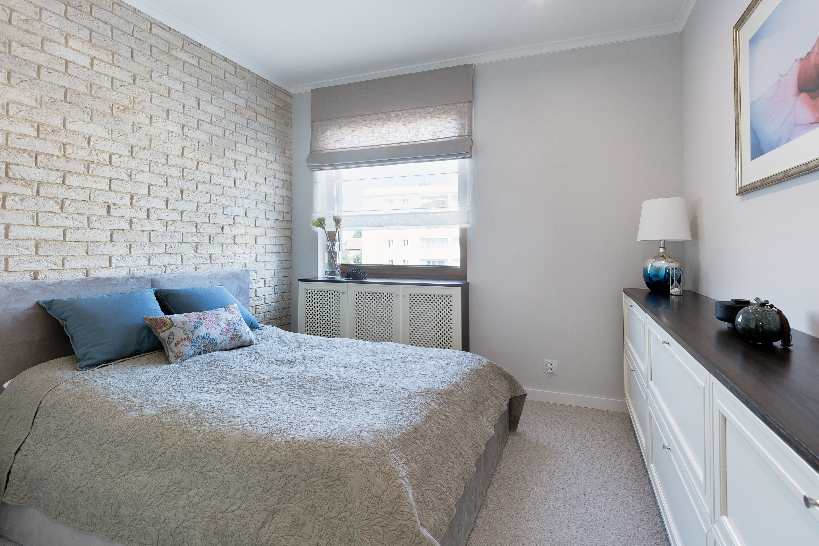 Jasna sypialnia z łóżkiem z beżową narzutą w roślinny wzór, niebieskimi poduszkami i ścianą z białej cegły dekoracyjnej, obok okna z roletą rzymską i białej komody z ciemnym blatem.