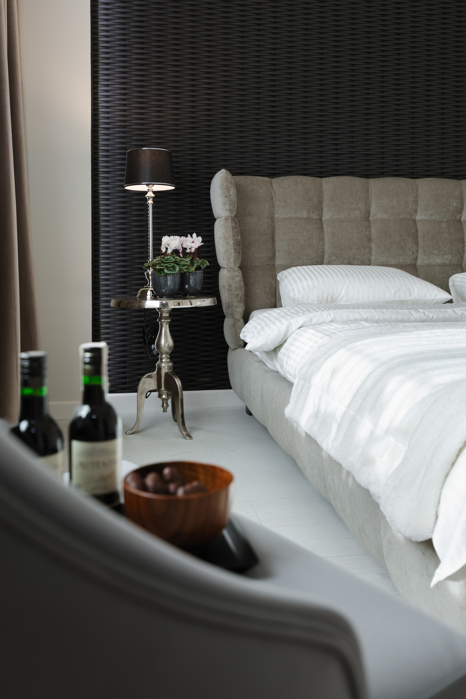 Nowoczesna sypialnia z szarym tapicerowanym łóżkiem, stolikiem nocnym ze srebrną lampą i kwiatami, oraz butelkami wina i miseczką czekoladek na pierwszym planie.