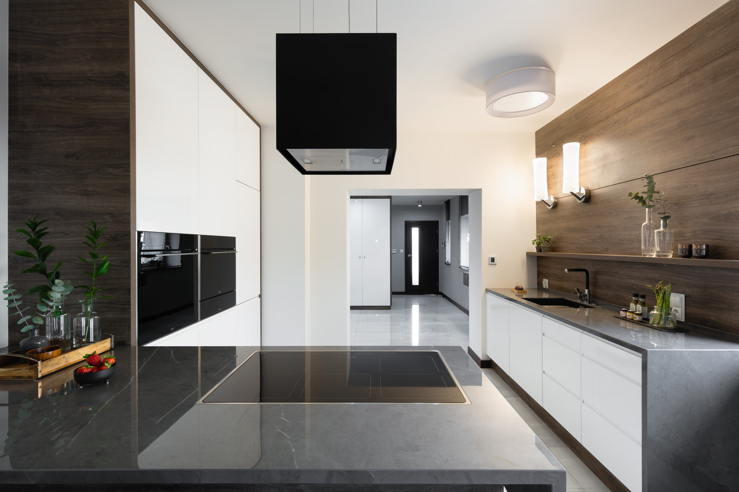 Nowoczesna kuchnia z wyspą z szarego marmuru, czarnym okapem i białymi szafkami, uzupełniona drewnianymi akcentami i dekoracyjnymi roślinami w szklanych wazonach.