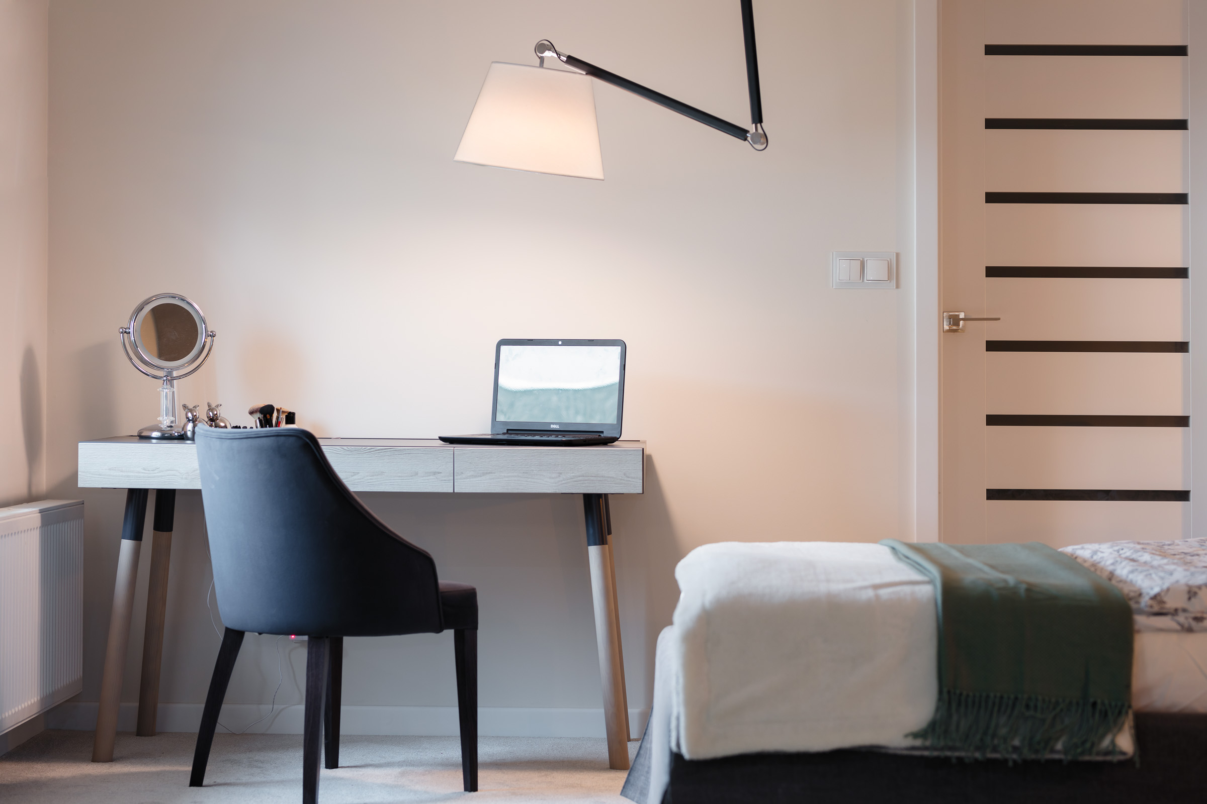 Nowoczesny pokój z biurkiem, laptopem, krzesłem, lustrem do makijażu i lampą wiszącą, minimalistyczny wystrój, białe ściany, drzwi z poziomymi przeszkleniami.