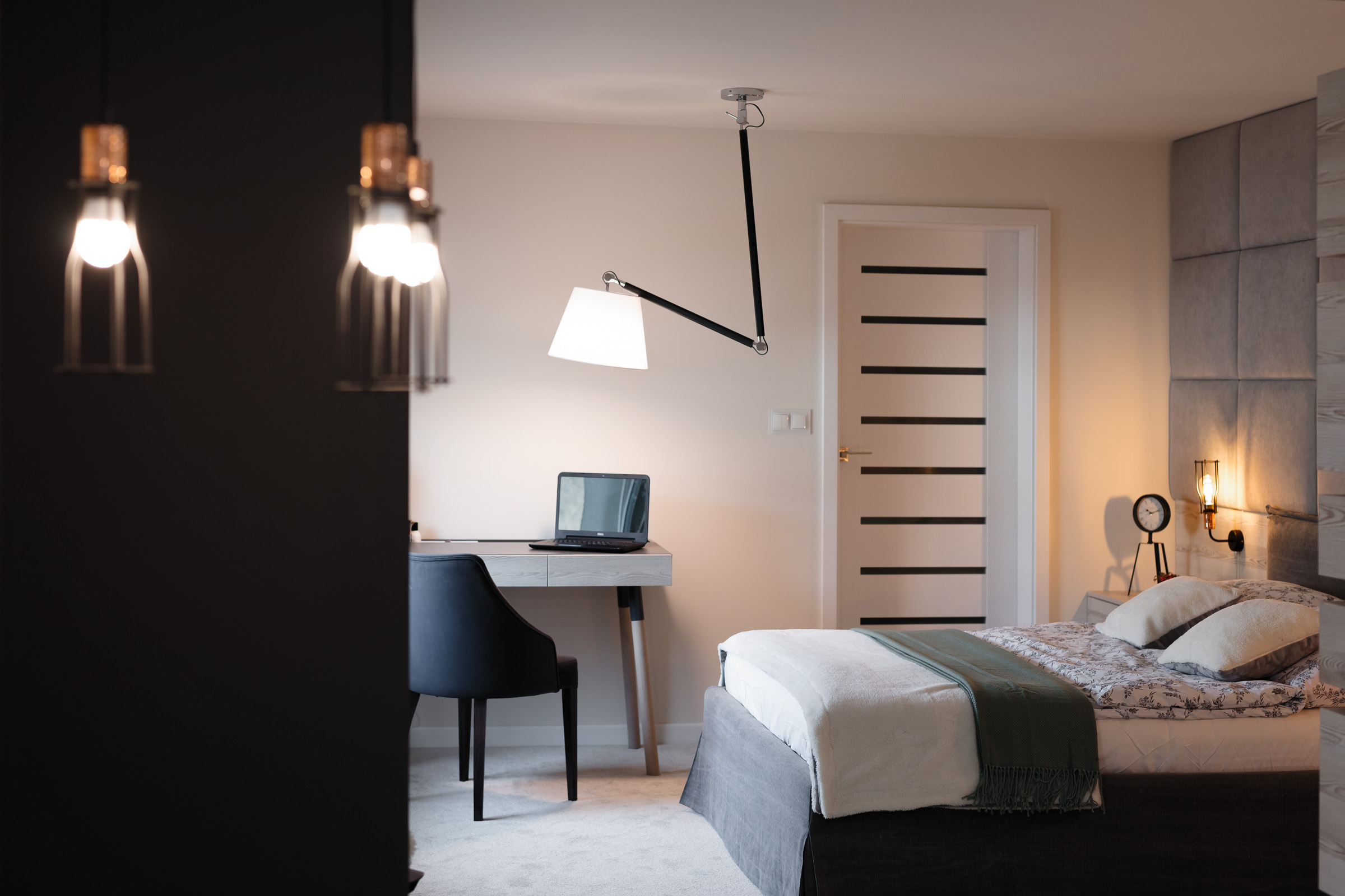 Nowoczesna sypialnia z biurkiem i lampą architektoniczną, stonowana kolorystyka, dekoracyjne oświetlenie nocne i panele tapicerowane za łóżkiem.