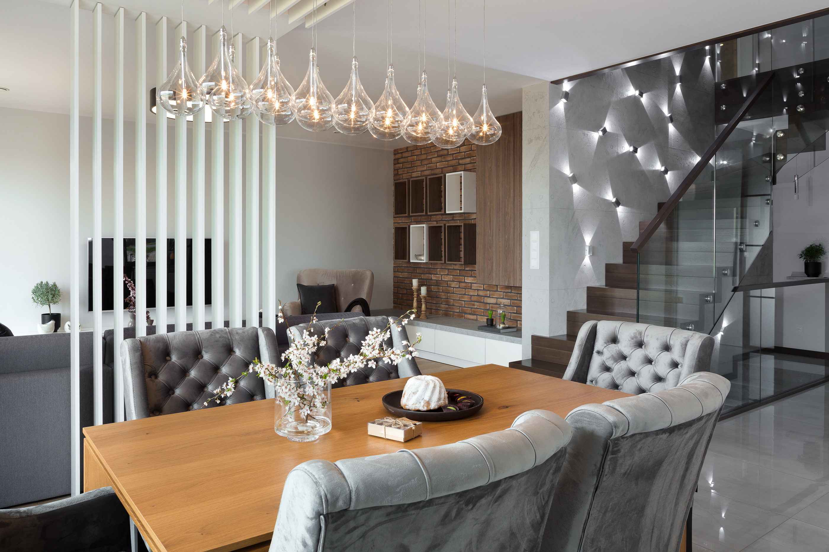 Nowoczesna jadalnia z drewnianym stołem, szarymi krzesłami z pikowaniem, dekoracyjnym oświetleniem sufitowym i szklanymi schodami w tle.