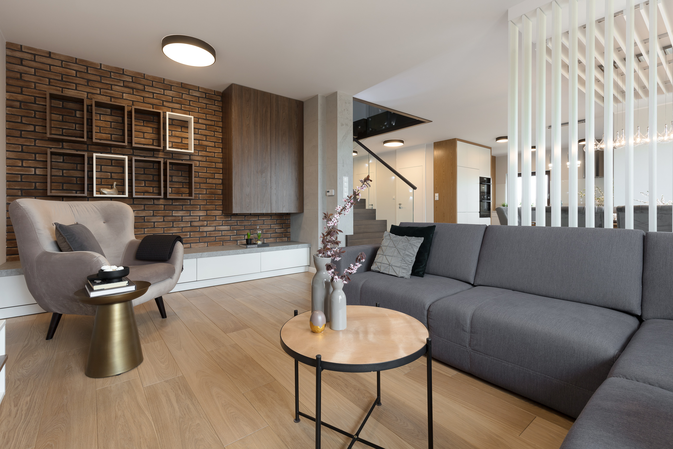 Nowoczesny salon z ceglaną ścianą, szarą sofą narożną, fotelem w odcieniach szarości i stolikami kawowymi o różnych kształtach, oświetlony ciepłym światłem.