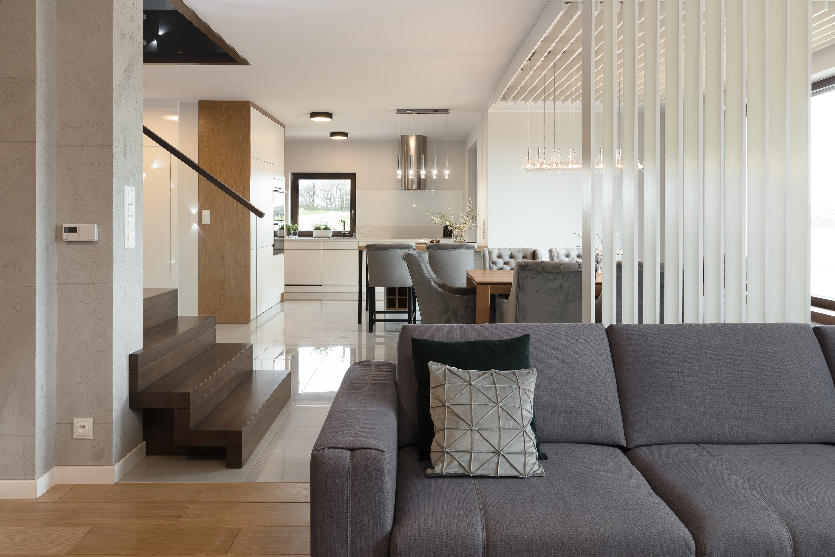 Przestronne wnętrze domu z szarym narożnikiem, drewnianymi schodami i białą kuchnią w tle, oświetlone wiszącymi lampami.