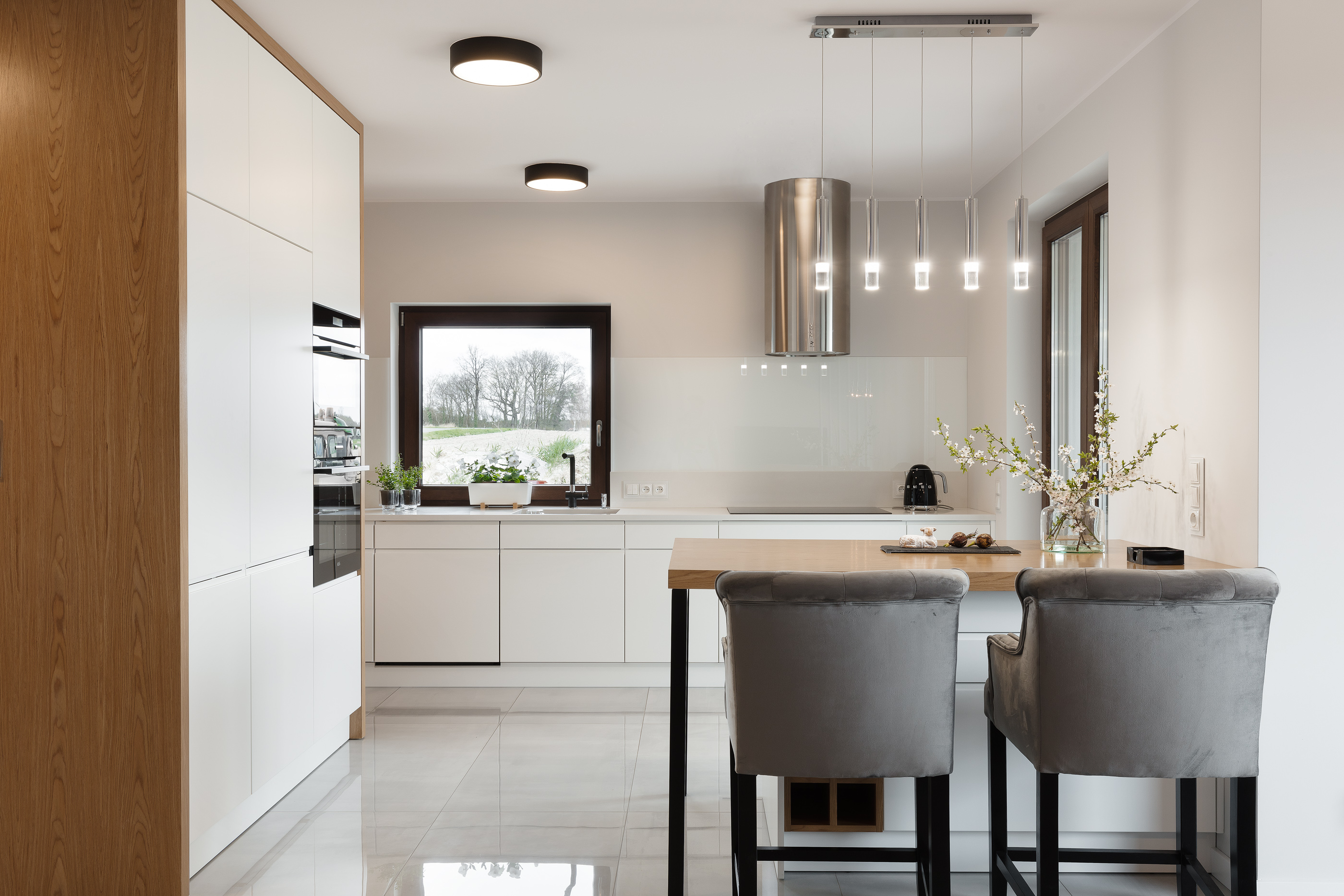 Nowoczesna, jasna kuchnia z wyspą i dwoma szarymi krzesłami barowymi. Na blacie wyspy wazon z kwitnącymi gałązkami, w tle okno z widokiem na ogród.