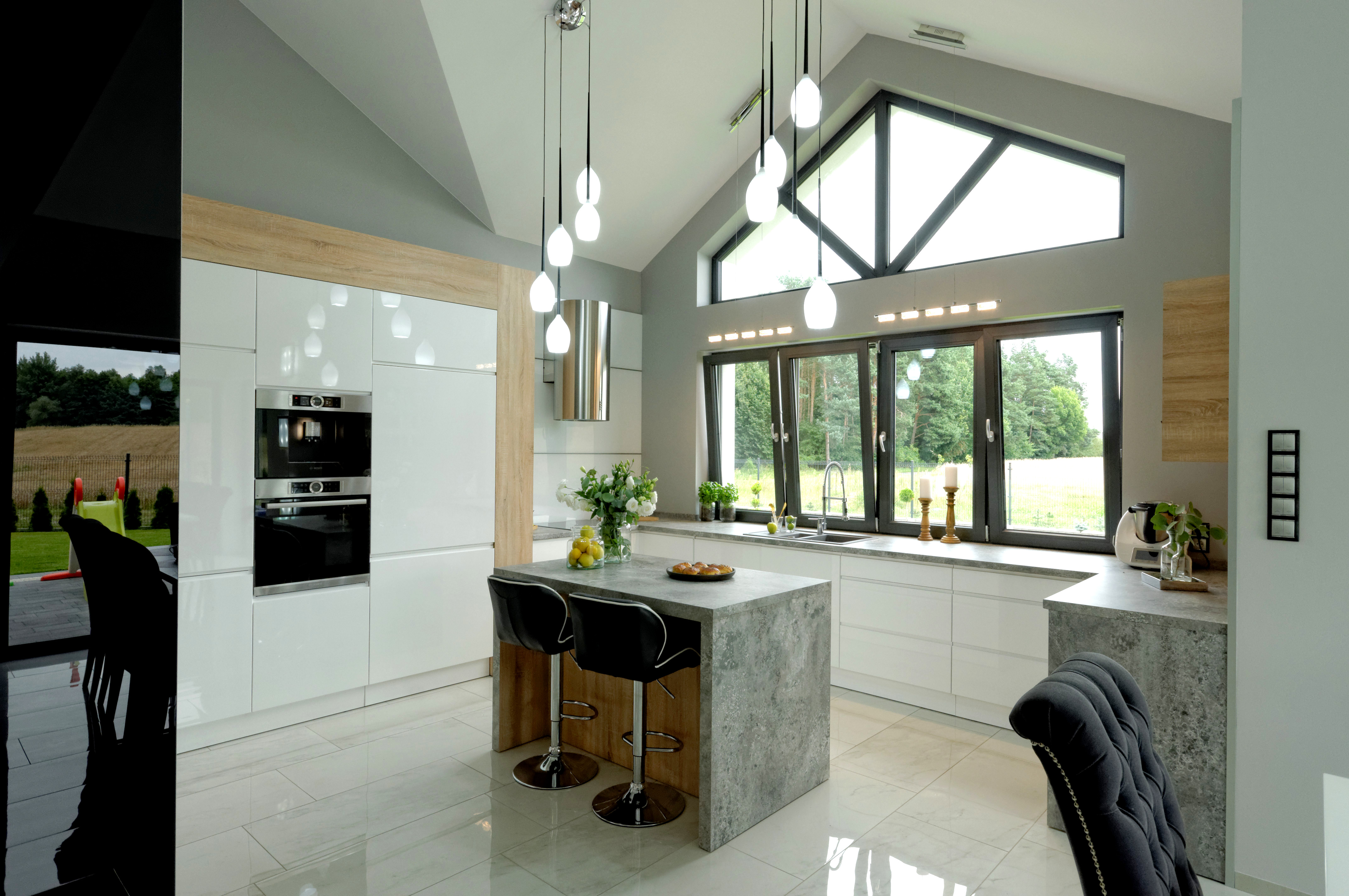 Nowoczesna kuchnia z wyspą i dużym oknem szczytowym, białe szafki z drewnianymi akcentami, marmurowa podłoga i wiszące lampy kuliste.
