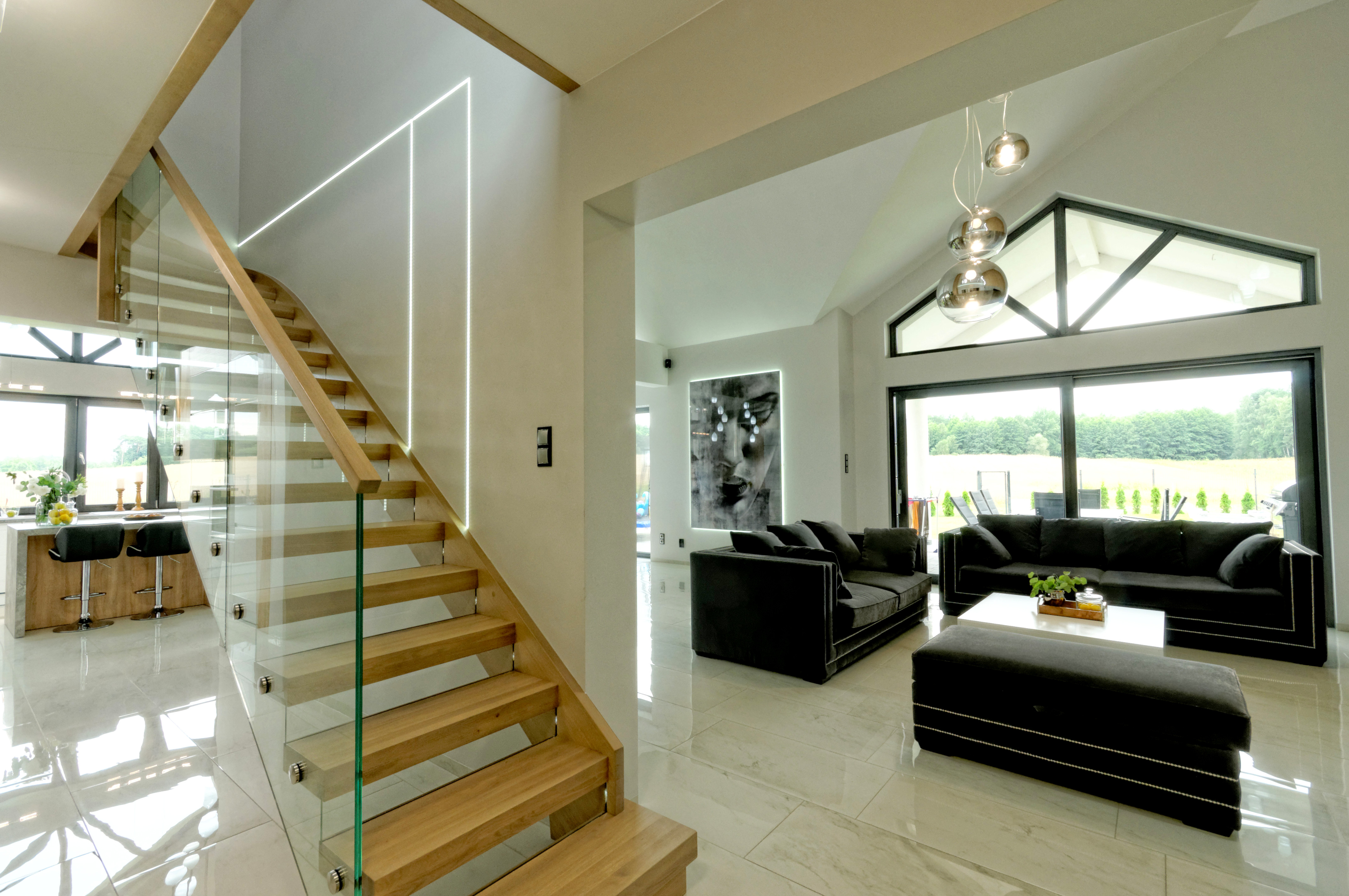 Nowoczesny salon z drewnianymi schodami z szklaną balustradą, oświetleniem LED na ścianie, czarnymi sofami i marmurową podłogą.
