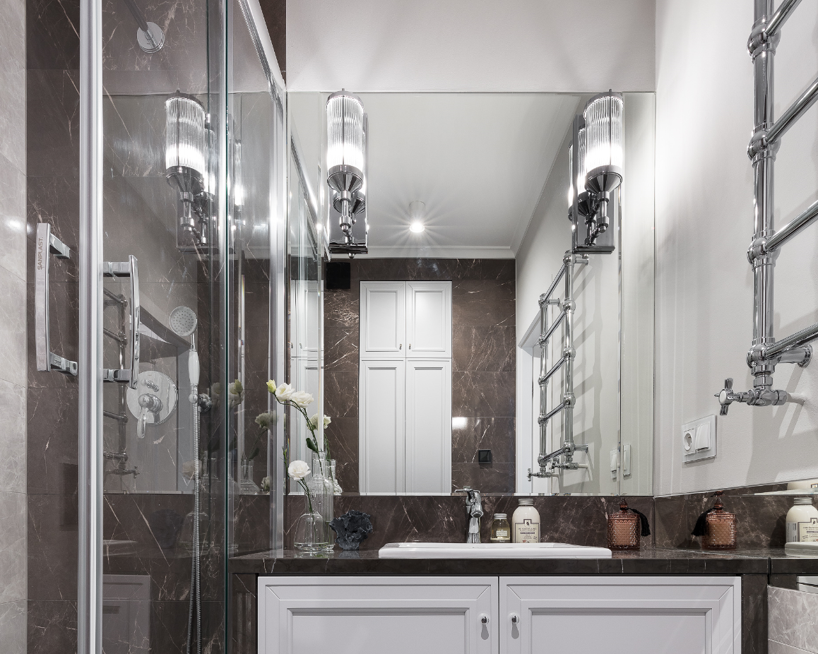 Nowoczesna łazienka z kabiną prysznicową, marmurowymi płytkami w odcieniach szarości, białymi szafkami pod umywalką i ozdobnymi lampami ściennymi z żłobionego szkła.