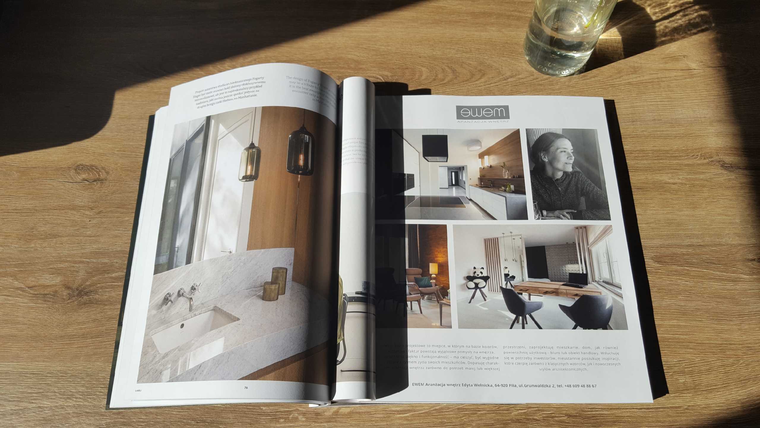 Otwarta gazeta na drewnianym stole z fotografiami wnętrz, w tym łazienki z marmurowym blatem i wiszącymi lampami, oraz kuchni i pokoju dziennego, obok szklanka z wodą.