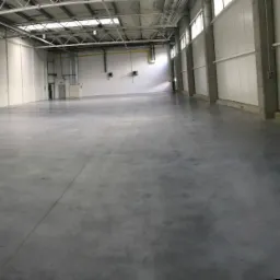 Wnętrze hali magazynowej z szarą, betonową podłogą, jasnymi ścianami z paneli i metalową konstrukcją dachu z oknami dachowymi, widok z poziomu podłogi.