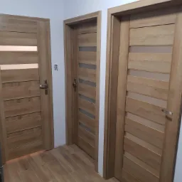 Trzy drewniane drzwi wewnętrzne z przeszkleniami w nowoczesnym mieszkaniu, widoczne na tle płytek podłogowych imitujących drewno.