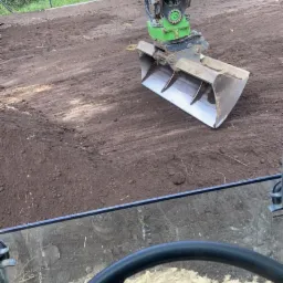 Widok z kabiny koparki na świeżo wyrównany teren z widocznym łyżką koparki i śladami opon w tle, zielone drzewa i ogrodzenie w oddali.