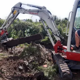 Mini koparka Takeuchi z hydrauliczną piłą do cięcia betonu, podczas pracy w wykopie, widoczne fragmenty gruzu i ziemi.