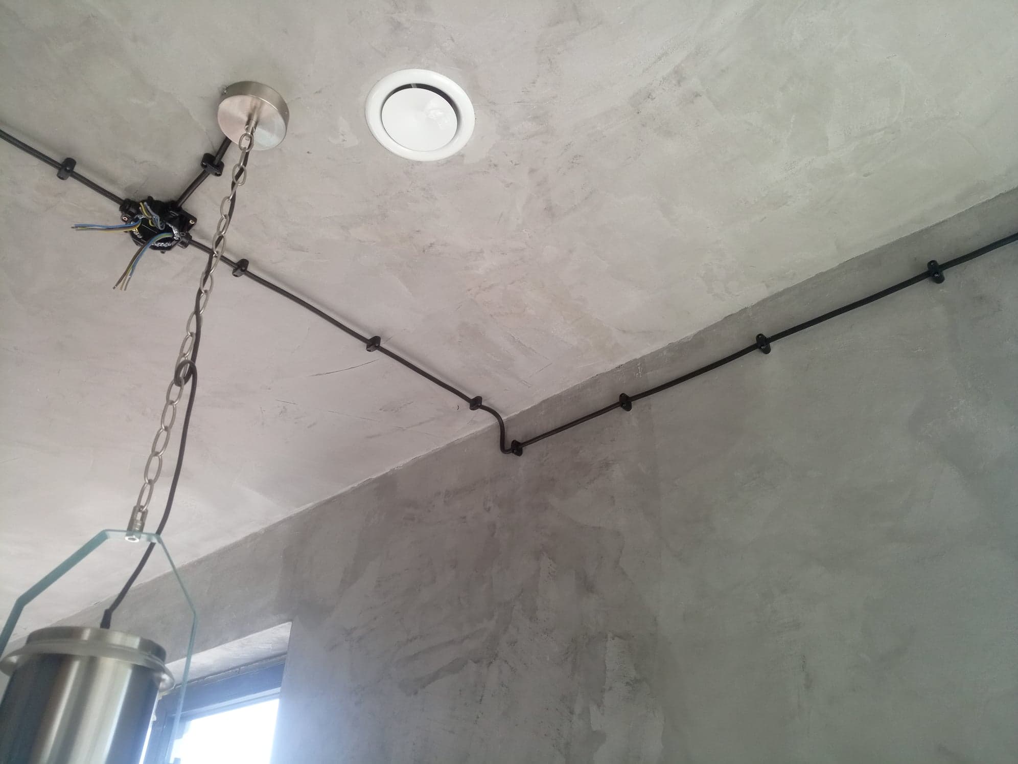 Betonowy sufit z widocznymi czarnymi przewodami elektrycznymi mocowanymi klamrami, centralnie umieszczony okrągły nawiewnik i podwieszona lampa z metalowym kloszem na łańcuchu. Widoczne...