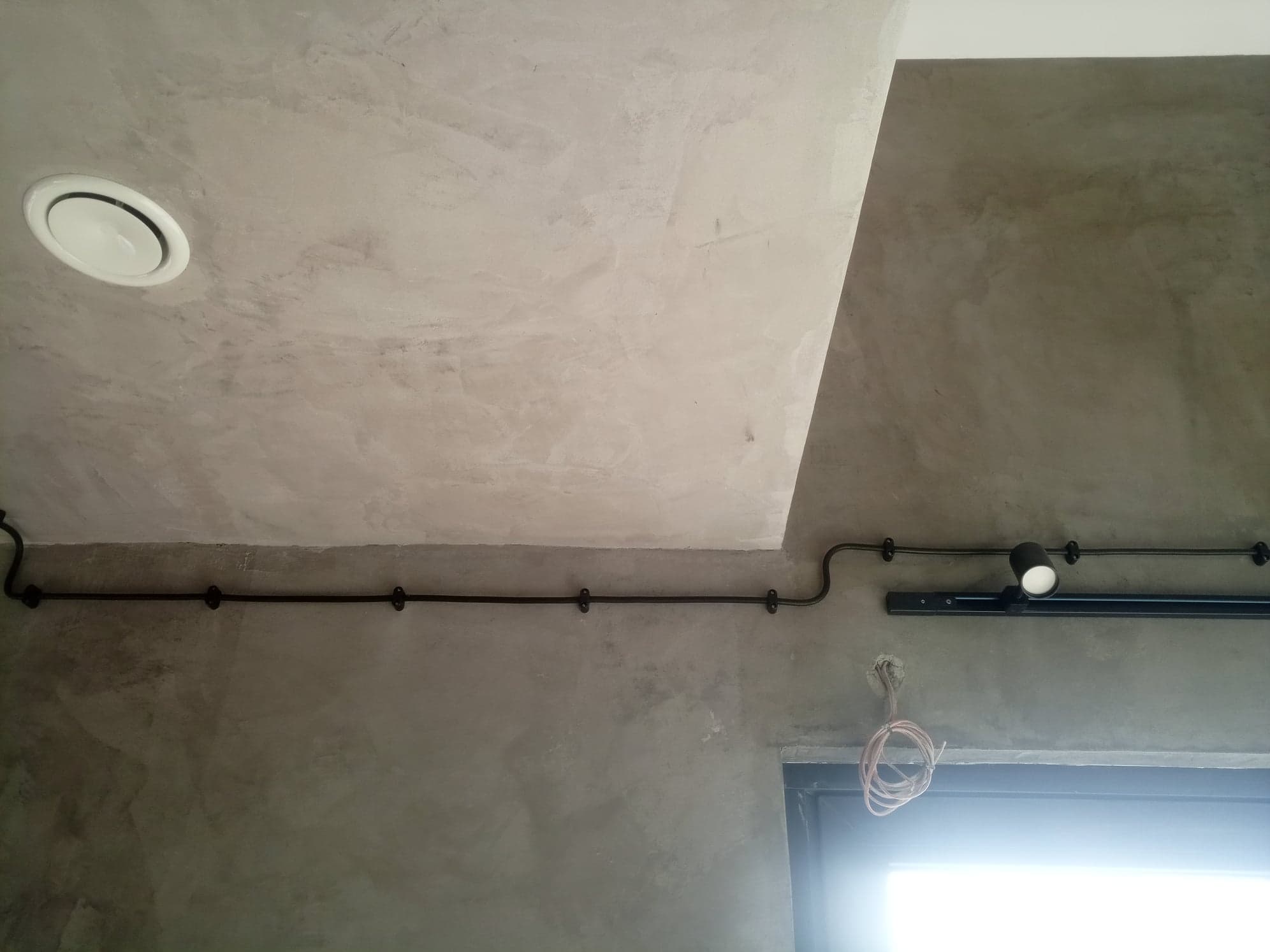 Betonowy sufit z widoczną instalacją elektryczną: przewody w czarnej rurce, reflektor na szynie i okrągły nawiewnik. W tle fragment okna lub drzwi.