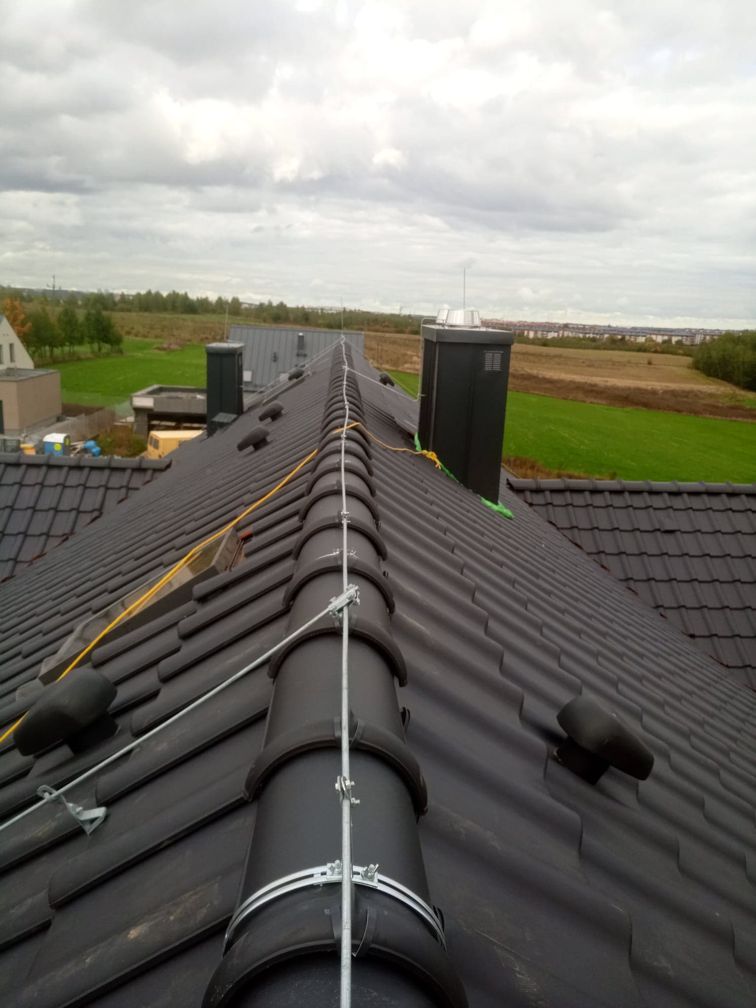 Widok z góry na dach z ciemnej dachówki ceramicznej z zamontowanym systemem odgromowym, na tle pochmurnego nieba i odległego krajobrazu pól.