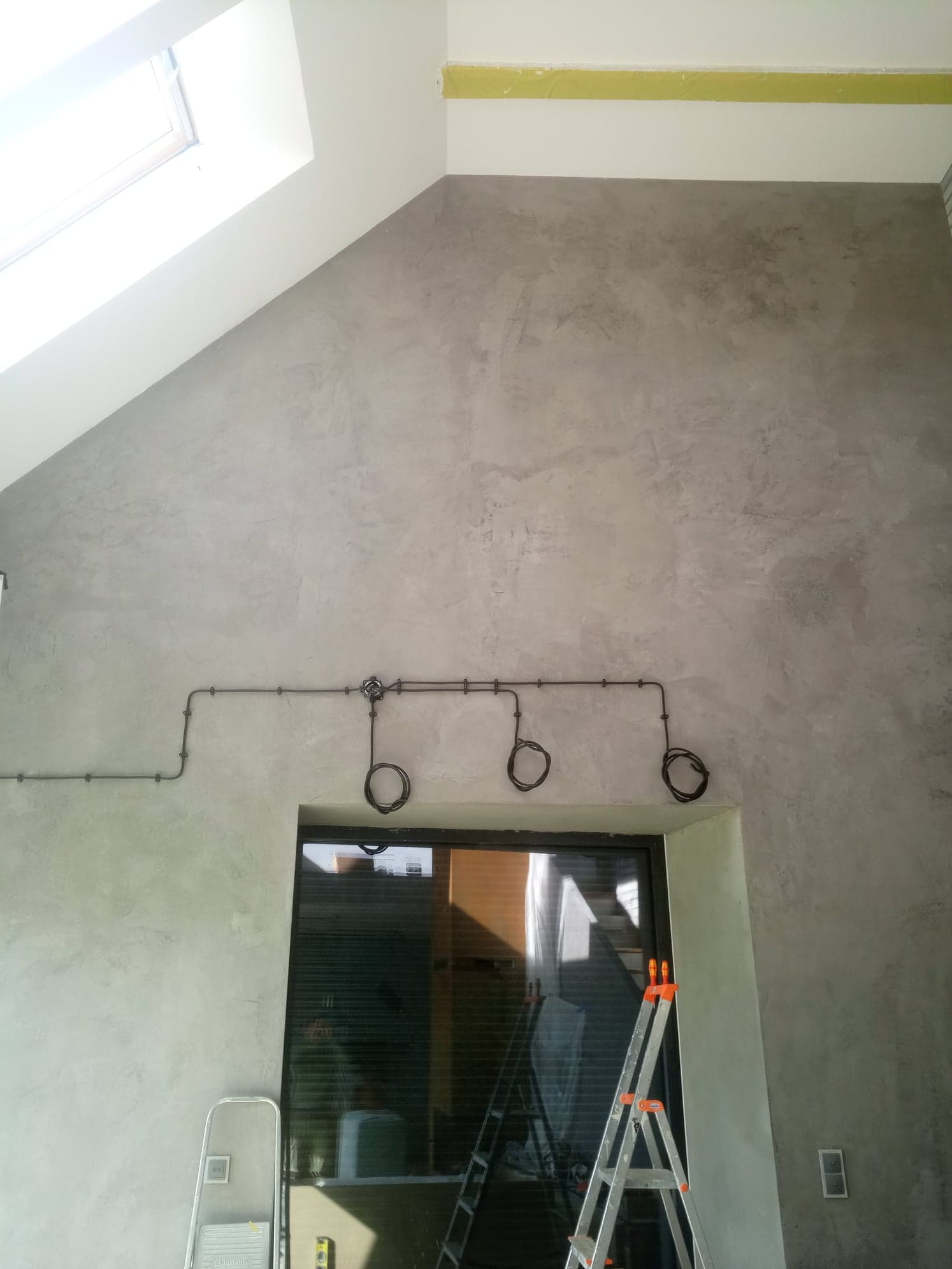 Szara ściana z wyeksponowanymi przewodami elektrycznymi w metalowych rurkach, widoczne schody w tle, okno dachowe i drabina.