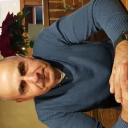 Mężczyzna w swetrze z zegarkiem na ręce siedzi przy stole, w tle dekoracja kwiatowa i ceglana ściana. Nietypowy kadr, oświetlenie ciepłe.