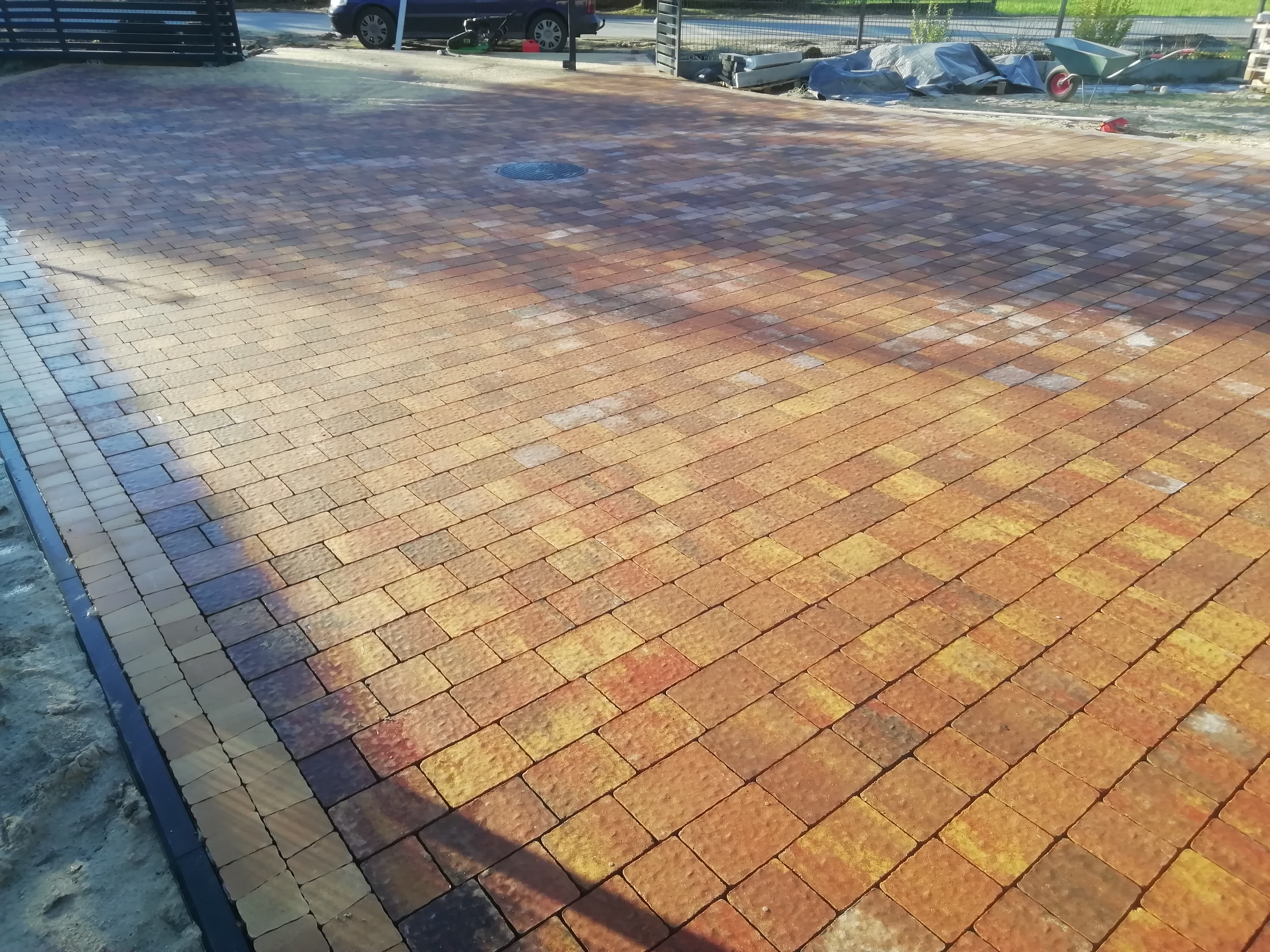 Ułożona kostka brukowa w odcieniach pomarańczy i żółci, widok z góry na dużą powierzchnię z widocznym włazem kanalizacyjnym, częściowo zacieniona z elementami ogrodzenia i pojazdu w tle.