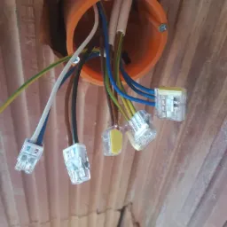 Pomarańczowa puszka instalacyjna z podłączonymi przewodami za pomocą przezroczystych złączek, widok od dołu na tle ceglanego stropu.