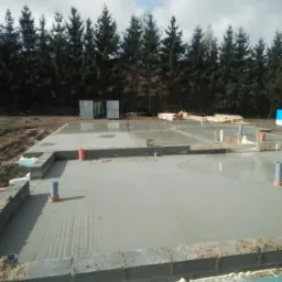 Świeżo wylany fundament domu jednorodzinnego z widoczną izolacją przeciwwilgociową, rurami instalacyjnymi i zarysowanymi śladami po szpachli na powierzchni betonu, w tle rząd drzew iglastych.