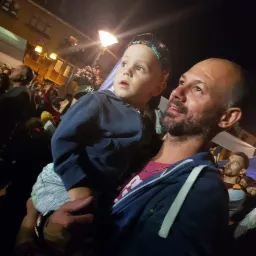 Ojciec trzyma syna na rękach, obaj patrzą w górę na tle nocnego miasta, widoczni inni ludzie i oświetlone budynki.