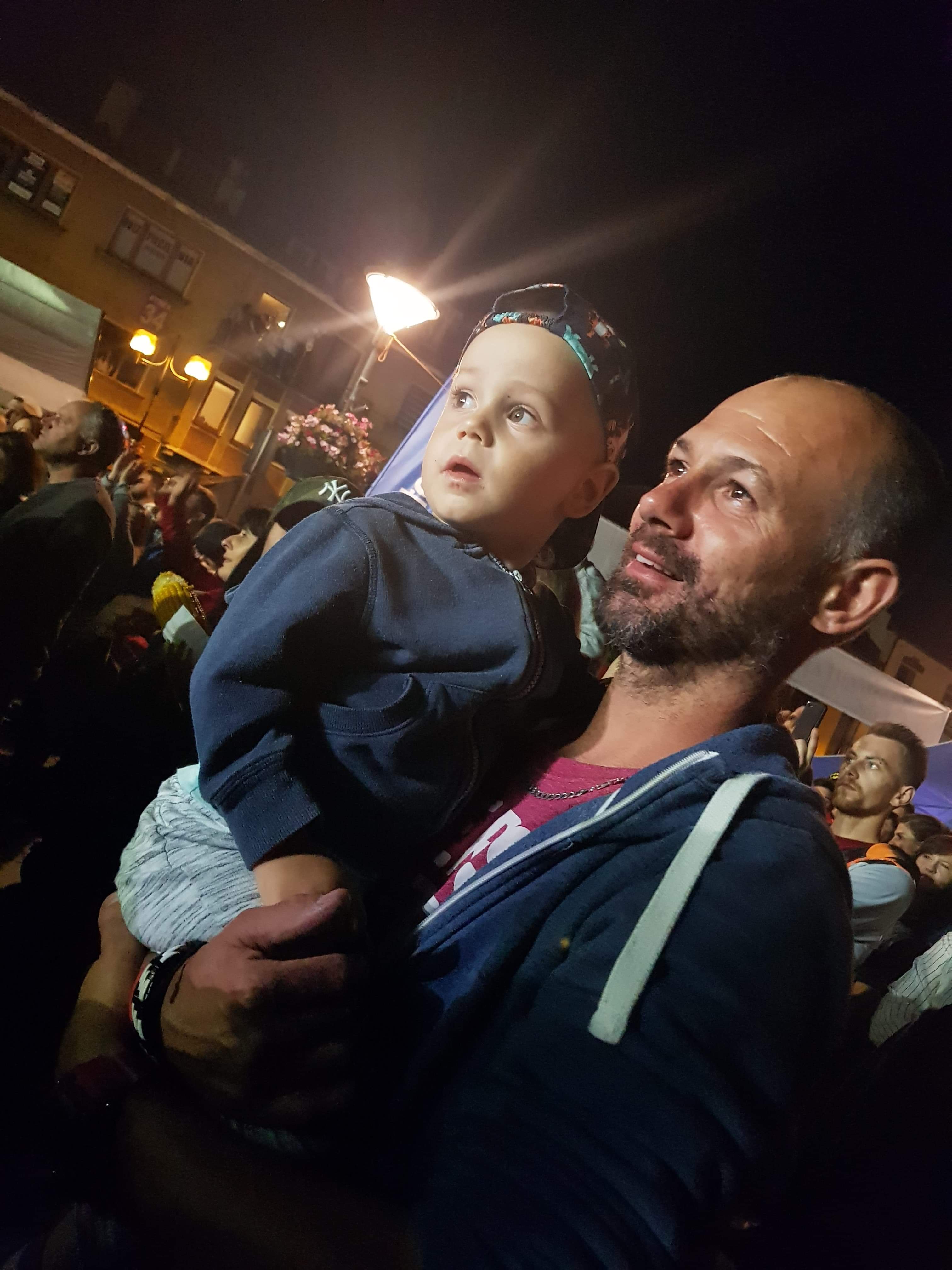 Ojciec trzyma syna na rękach, obaj patrzą w górę na tle nocnego miasta, widoczni inni ludzie i oświetlone budynki.