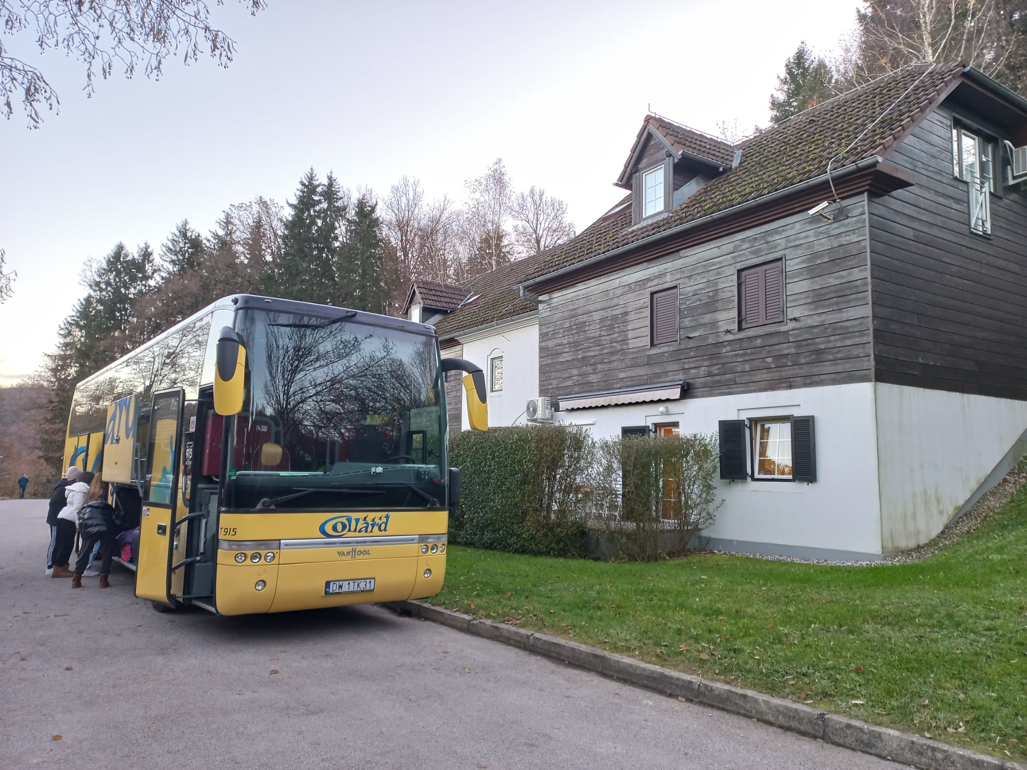 Żółty autokar turystyczny z otwartymi drzwiami, pasażerowie wsiadają, na tle budynku z drewnianą elewacją i zielenią.