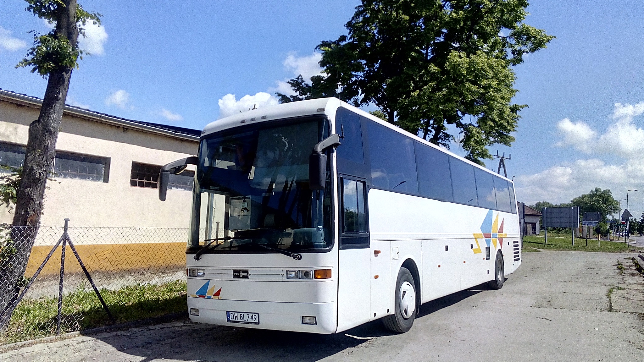 Biały autokar Van Hool z kolorowymi akcentami stoi na parkingu, widoczny numer rejestracyjny DW 8L749, w tle budynek i drzewa.