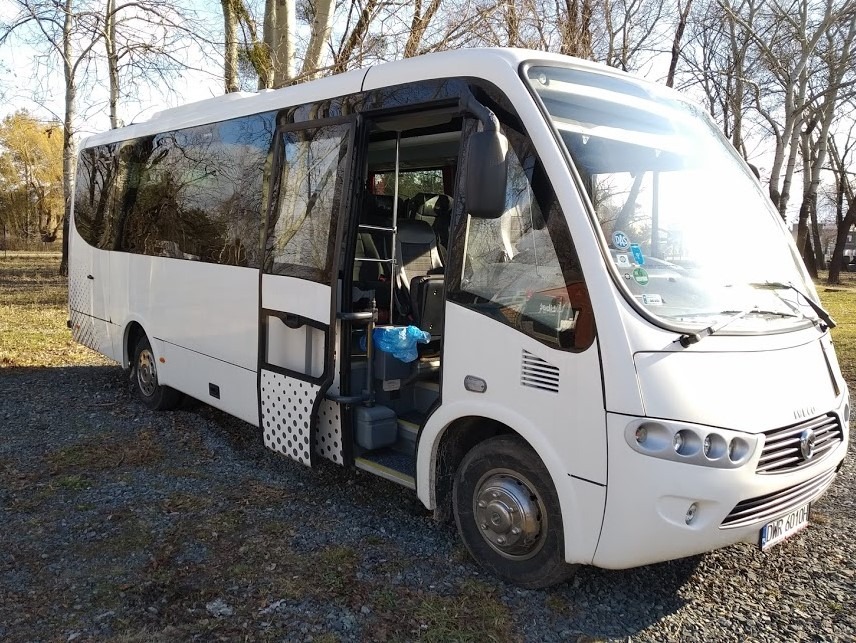 Biały minibus Iveco z otwartymi drzwiami, widoczne wnętrze, na szarym żwirowym podłożu, w tle drzewa bez liści.