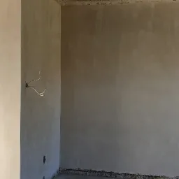 Surowe wnętrze w trakcie prac wykończeniowych, świeżo położone tynki na ścianach, widoczne wystające kable elektryczne, okno z widokiem na zieleń na zewnątrz.