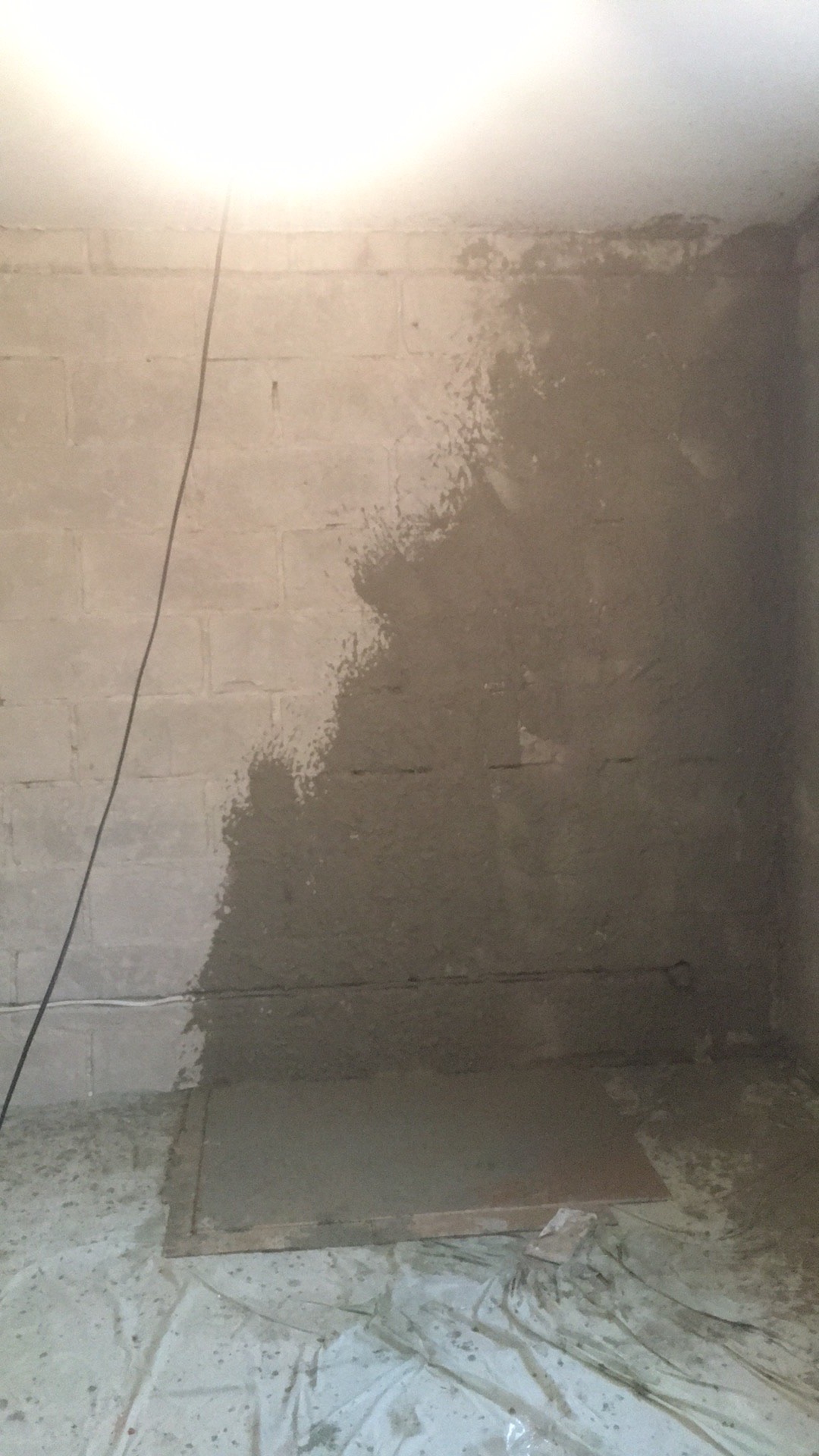 Fragment ściany z bloków, częściowo pokrytej świeżo nałożoną warstwą szarego tynku; podłoga zabezpieczona folią malarską.