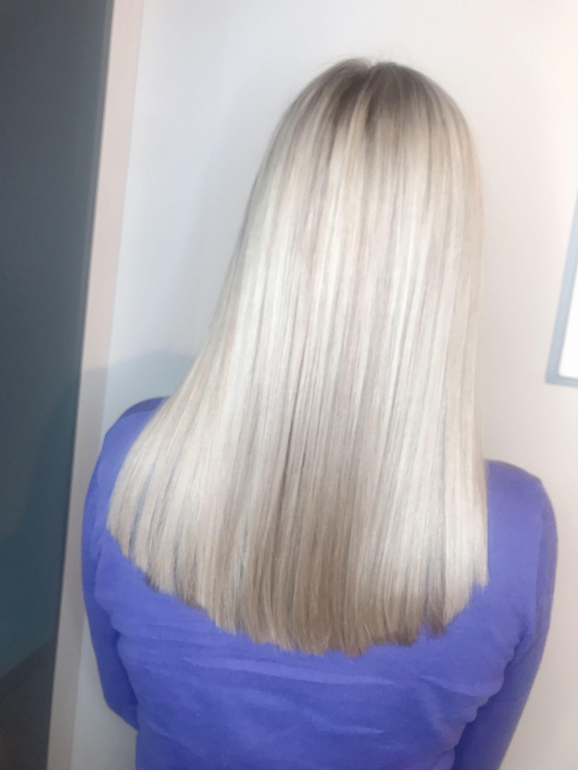 Tył głowy z długimi, prostymi, blond włosami z ciemniejszymi odrostami, na tle ściany w odcieniach szarości i bieli. Osoba ubrana w fioletową bluzkę.